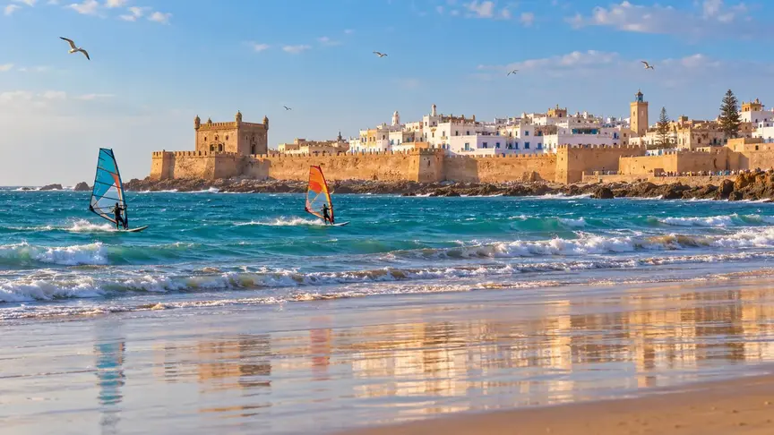 Essaouira-inspired illustration of windsurfers on Atlantic waves beside historic ramparts and whitewashed buildings.